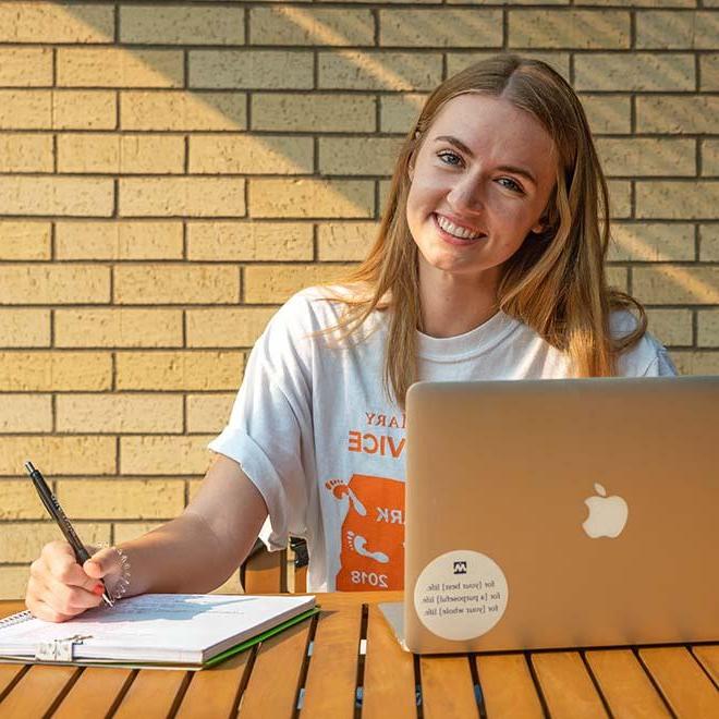 微笑的学生在外面，笔记本电脑和笔记本在她面前的桌子上